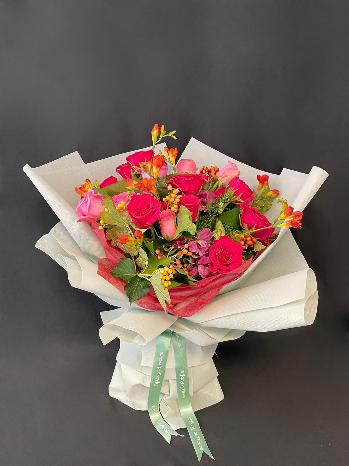 Elegant wrapped bouquet with hot pink roses, orange freesia, hypericum berries, alstroemeria, and ivy, finished with signature Tiffany et Fleurs ribbons — perfect for hospital visits or heartfelt gestures.