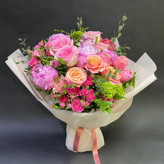 A soft-hued Mother's Day bouquet in pastel pinks and peaches, including roses and peonies, elegantly presented in a paper wrap with a pink ribbon.