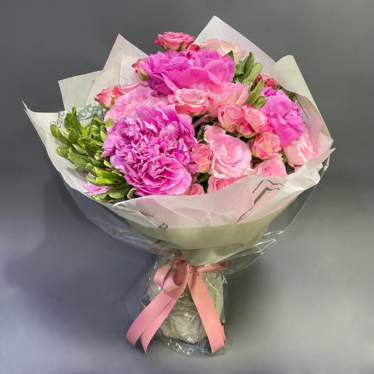 A vivid Mother's Day bouquet with lush pink peonies, delicate roses, and fresh greenery, wrapped in translucent paper with a satin bow.