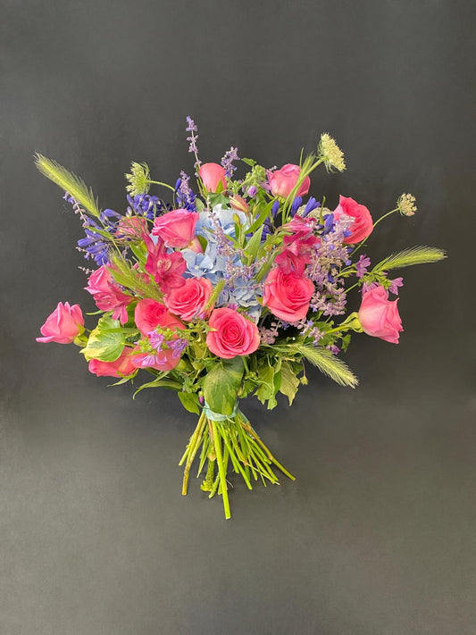 Hand-tied bouquet featuring vibrant pink roses, blue hydrangea, purple delphinium, lavender sprigs, and seasonal greenery — designed to uplift and brighten any recovery setting.